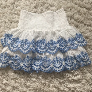 Vit kjol med blå broderi från Lindex - Supersöt vit kortkjol från Lindex med strech i midjan och flera lager volanger. Kjolen har detaljerade blå broderier längs kanterna som ger en fräsch och somrig vibe. Perfekt för dig som gillar unika detaljer och vill sticka ut lite extra. Inte använd en endaste gång.
