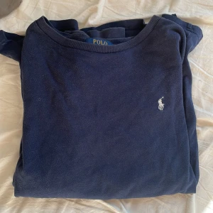 Mörkblå långärmad tröja Polo Ralph Lauren - Mörkblå långärmad tröja från Polo Ralph Lauren med klassisk rund hals och broderad vit logga på bröstet. Tillverkad i mjuk bomull, perfekt för en chill och stilren look. Passar dig som gillar enkel men snygg design.
