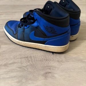 Nike Air Jordan 1 blå och svarta - Säljer ett par Nike Air Jordan 1 i blått och svart skinn med klassisk high-top siluett. Skorna har svarta snören, perforerad tåbox och vit sula. Ikoniska Jordan Wings-loggan på sidan och Jumpman på plösen. Perfekta för dig som gillar streetwear och sneakers med attityd.