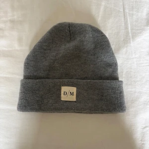 Grå mössa - En stilren grå mössa med uppvikt kant och en liten vit logotyp framtill. Perfekt för kalla dagar och enkel att matcha med olika outfits. Tillverkad i mjukt material som känns skönt mot huden.