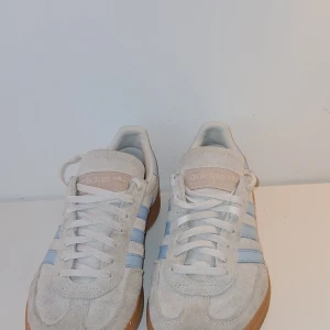 Adidas Handball Spezial ljusblå/vita - Säljer nu mina Adidas Handball Spezial sneakers i mocka med beige bas, ljusblå ränder och detaljer samt klassisk gummisula. Skorna har snörning och rund tå, perfekt för dig som gillar ett par klassiska skor.  Säljer p.g.a att jag växte ur dem lite för fort, bra skick.🤍💙               Nypris: 1300 kr