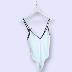 Vit baddräkt med svarta detaljer från Nelly - Supersnygg vit baddräkt från Nelly med tunna svarta axelband och v-ringning. Stilren design med svarta kanter som ger en clean kontrast. Perfekt för stranden eller poolen, och sitter bekvämt med stretchigt material. Aldrig använd.