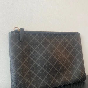 Svart kuvertväska från By Malene Birger - Stilren svart kuvertväska från By Malene Birger med rutigt mönster och guldfärgad dragkedja. Väskan är tillverkad i syntetmaterial med struktur och har en enkel, rektangulär form. Perfekt att bära till det mesta när du vill ha med det viktigaste. Saknar bandet till.