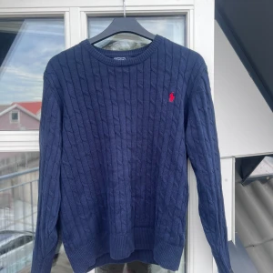 Marinblå kabelstickad tröja Polo Ralph Lauren - Kabelstickad marinblå tröja från Polo Ralph Lauren med klassisk rund hals och röd broderad logga på bröstet. Långärmad modell i mjuk bomull, perfekt för lager-på-lager och snygg till jeans eller chinos.