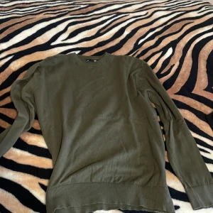 Olivgrön tröja från lager 157 - Enkel och stilren olivgrön tröja från 157 i storlek L. Tröjan har rund halsringning, långa ärmar och breda ribbade muddar vid ärmslut och nederkant. Perfekt för lager-på-lager och passar till det mesta i garderoben.