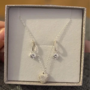 SILVERSET SNÖ halsband & örhängen - Supersött smyckeset från SNÖ med ett halsband i silverfärg och hjärtformat hänge täckt av små gnistrande stenar. Matchande örhängen med glittrande stenar. Perfekt present, levereras i glittrig ask med rosett.