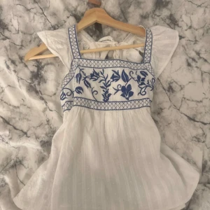 Vit blus med blå broderi och volang - Supersöt vit blus med blå broderade blommor och rutmönster upptill. Blusen har volangärmar, fyrkantig ringning och knytning i ryggen. Lätt och luftig i bomull, perfekt för varma dagar när du vill ha en romantisk vibe.