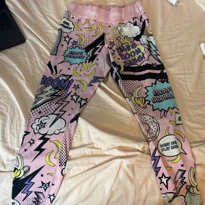 Färgglada leggings från Stronger - Säljer ett par supercoola leggings från Stronger med serietidningsinspirerat tryck i rosa, gult, svart och blått. De har hög midja och är tillverkade i stretchigt material som passar perfekt till träning. Massor av detaljer som blixtar, stjärnor och textbubblor.