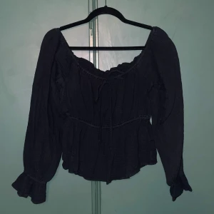 Svart off shoulder blus med volang från NLY Trend - Supersöt svart off shoulder blus från NLY Trend med långa ärmar och volangdetaljer vid ärmslut och halsringning. Blusen har knytning framtill och är tillverkad i ett mjukt, strukturerat bomullstyg.  
