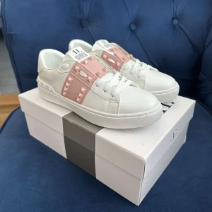 Valentino Rockstud sneakers vit/rosa - Snygga Valentino Rockstud sneakers i vitt skinn med ljusrosa detaljer och ikoniska nitar. Klassisk rund tå, platt sula och vita snören. Perfekta för dig som vill ha en lyxig och trendig look med en touch av rosa och coola metalldetaljer.
