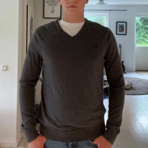 Merino tröja från Fred Perry - Snygg mörkgrå v-ringad tröja från Fred Perry med diskret logga på bröstet. Tröjan har lång ärm och ribbade muddar vid ärmslut och nederkant. Perfekt att slänga på över en t-shirt för en clean och stilren look.