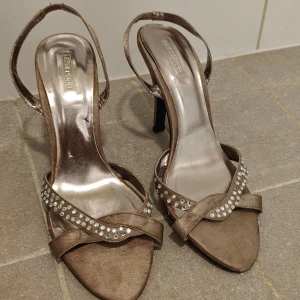 Högklackade skor beige strl 40 - Snygga beige klackar från La Strada med öppen tå och smala remmar dekorerade med stenar. Elegant design, perfekta för fest eller bröllop.Materialet har en satinliknande finish som ger extra lyxig känsla. Ej slitet på undersidan.