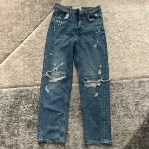 Blå 90's Straight jeans från H&M - Säljer ett par blå 90's Straight jeans från H&M med slitna detaljer och hål på knäna. Jeansen har rak passform, hög midja och klassisk femficksdesign. Perfekta för en avslappnad och trendig look.