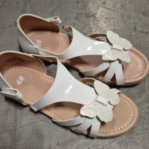 Sandaler - Söta vita sandaler från H&M med glittriga fjärilar framtill. Skorna har flera remmar, öppen tå och justerbar rem runt hälen. Insidan är ljusbeige och baksidan har en lätt skimrande yta. Perfekta för varma dagar och en extra touch av glitter.