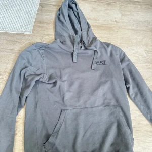 Grå hoodie från EA7 Emporio Armani - Snygg grå hoodie från EA7 Emporio Armani med klassisk huva och snörning. Tröjan har en stor magficka och diskret logga på bröstet. Perfekt för en chill och stilren look. Tillverkad i mjuk bomull för maximal komfort.