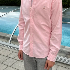 Rosa skjorta från Ralph Lauren Slim Fit - Säljer en stilren rosa skjorta från Ralph Lauren i slim fit-modell. Skjortan har klassisk krage, knäppning framtill och det ikoniska broderade polospelar-loggan på bröstet. Tillverkad i mjuk bomull och med långa ärmar. Perfekt för dig som gillar en clean och fräsch look.