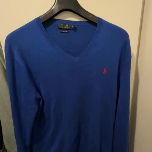 Ralph Lauren v-ringad sweatshirt - Säljer en Ralph Lauren V-ringad sweatshirt i blått, med röd broderad logga.  Tillverkad i exklusiv Pima cotton - extra mjuk och följsam med lyxig känsla.  En stilren och mångsidig tröja som passar både till vardags och lite mer uppklätt.  Storlek: M (normal passform)   Skick: Väldigt fint – inga fläckar, hål eller slitage    Färg: Blå med röd broderad hästlogga  Material: Pima cotton – mjukt, hållbart och högkvalitativt
