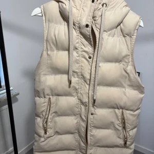 Beige dunväst från Zara med huva - Säljer en mellan lång beige dunväst från Zara i storlek XS. Västen har en quiltad design, dragkedja och tryckknappar framtill, två fickor med dragkedja och en stor huva med snörning. Perfekt att slänga över en hoodie för en chill look.