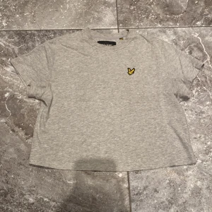 Grå t-shirt från Lyle & Scott - Fåtals gånger används, nyskick! Snygg grå t-shirt från Lyle & Scott i 100% ekologisk bomull. Klassisk passform med korta ärmar och rund hals. På bröstet sitter den ikoniska gula örnloggan som ger en clean och stilren vibe. Perfekt till jeans eller shorts för en chill look.