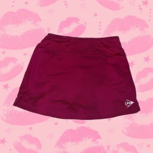 Rosa sportkjol från Dunlop - Supersnygg rosa sportkjol med shorts / skort från Dunlop med elastisk midja och diskret logga framtill. Kjolen är kort och tillverkad i ett lätt och stretchigt syntetmaterial som passar perfekt för tennis eller träning. Enkel och stilren design som ger en sportig vibe. Ungefärliga mått - 28 cm över midjebandet - 31 cm, midjebandet är lite stretchigt från topp till botten
