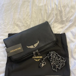 Svart axelväska Zadig & Voltaire - Snygg svart axelväska från Zadig & Voltaire i skinn med silverfärgad kedjerem och dragkedja. Väskan har ett coolt emblem med glittriga vingar framtill och stilren design. Perfekt för dig som vill ha en edgy och trendig accessoar.