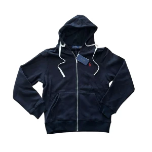 Ny Ralph Lauren Hoodie - Svart hoodie från Polo Ralph Lauren med dragkedja, vit snörning i huvan och klassisk röd logga på bröstet. Tillverkad i mjuk bomullsmix, perfekt för chill dagar. Fickor framtill och ribbade muddar för extra komfort. Jag anser att den är ganska liten för att vara storlek L så skriv så skickar jag måtten :)