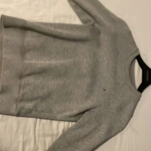 Grå sweatshirt från Tommy Hilfiger - Säljer en stilren grå sweatshirt från Tommy Hilfiger. Tröjan har en klassisk rund halsringning och långa ärmar. Perfekt för en avslappnad look. Den har en liten logotyp på bröstet och ribbade muddar vid ärmslut och nederkant.