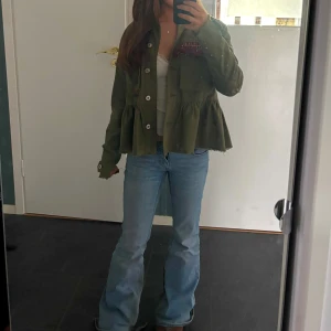 Grön jacka med volangdetaljer - Snygg grön jacka med volangdetaljer och knappar framtill. Jackan har en unik design med broderade detaljer i rött på bröstet. Perfekt för att ge en stilfull touch till din outfit. Sista bilden visar färgen bäst! ❤️