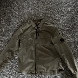 Olivgrön overshirt från C.P. Company - Snygg olivgrön overshirt från C.P. Company med dragkedja och två bröstfickor. Jackan har en klassisk krage och en rund detalj på ärmen. Perfekt för en stilren look.