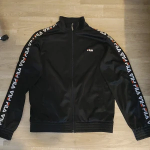 Svart träningsjacka från FILA - Snygg svart träningsjacka från FILA med dragkedja och logotyp längs ärmarna. Perfekt för en sportig look. Jackan har en hög krage och ribbade muddar för extra komfort.