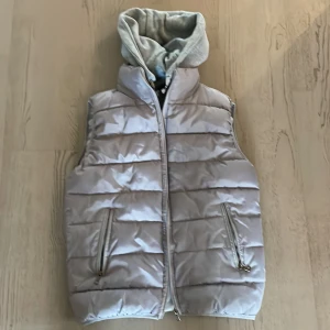 Grå quiltad väst med huva - Snygg grå quiltad väst med dragkedja och Västen har praktiska fickor med dragkedja framtill. Funkar från tidig vår till sen höst. Knappt använd alls. Från all the men.