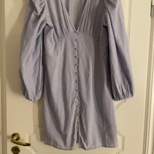 Ljus lilla klänning från Vero Moda - Säljer en ljus lilla  klänning från Vero Moda med v-ringning och knappar framtill. Klänningen har långa ärmar och ett broderat mönster. Perfekt för vår och sommar!