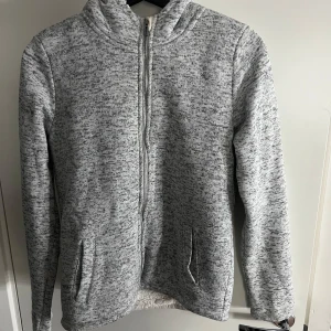 Grå fleecejacka från L.O.G.G. - Mysig grå fleecejacka från L.O.G.G. med dragkedja framtill och två fickor. Perfekt för kyliga dagar med sin mjuka insida. Jackan har en hög krage och långa ärmar för extra värme.