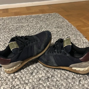 Valentino Garavani sneakers Rockrunners - Snygga Valentino Garavani sneakers i mörkblått med vita och vinröda detaljer. Skorna har en unik design med nitar på sulan och snörning framtill. Perfekta för en stilren look. Kan sänka priset. Man kan se hur de sitter på på ett av mina andra inlägg.
