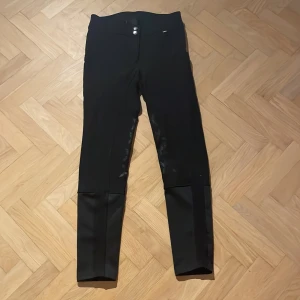 Svart ridbyxor från CRW, Hööks - Snygga halvskodda ridbyxor med justerbar midja, Storlek 36 och passar en 155 cm lång. Har fickor på båda byxbenen. Säljes pga för små