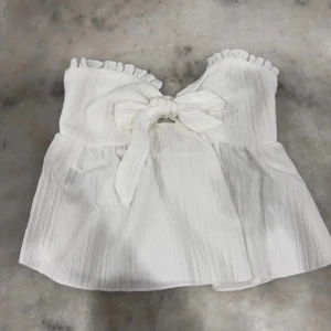 Vit axelbandslös topp med knyt - Säljer en söt vit axelbandslös topp med volangdetaljer och knyt framtill. Toppen har en smockad baksida för bättre passform och är perfekt för varma sommardagar. Passar bra till både jeans och kjol. 🌸