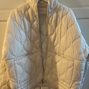 Beige quiltad jacka - Säljer en beige quiltad jacka med dragkedja och hög krage. Jackan har långa ärmar och två stora fickor framtill. Perfekt för kyligare dagar med sin mysiga och stilrena design. Aldrig använt! Helt ny