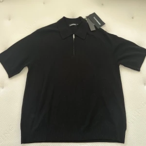 Svart merinoullströja från J.Lindeberg - Snygg svart tröja från J.Lindeberg i mjuk merinoull. Tröjan har en dragkedja framtill och korta ärmar, perfekt för en stilren look. Passar bra till både jeans och chinos. Ny pris 2200kr