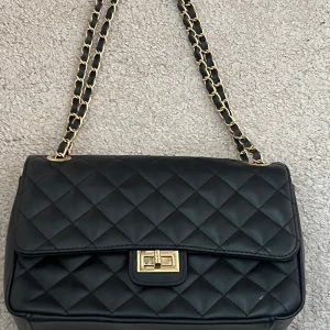 Svart quiltad axelväska från Isabella Rhea - Snygg svart quiltad axelväska från Isabella Rhea med guldfärgade kedjedetaljer. Väskan har en klassisk design med en praktisk låsdetalj framtill. Perfekt för att ge en elegant touch till din outfit.