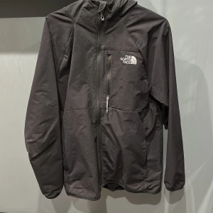 Svart vindjacka från The North Face - Säljer en svart vindjacka från The North Face med dragkedja och huva. Jackan har en enkel design med logotyp på bröstet och är perfekt för blåsiga dagar. Den har långa ärmar och praktiska fickor framtill.