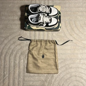 Svartvita sneakers från BAPE - Snygga svartvita sneakers från BAPE med klassisk design och snörning. Skorna kommer i en cool camouflagemönstrad låda och med en beige påse med BAPE-logga. Perfekta för dig som vill ha en stilren och trendig look.
