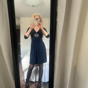 Mörkblå klänning från Your Sixth Sense - Elegant mörkblå klänning med v-ringning och en dekorativ detalj i midjan. Klänningen har trekvartsärmar och är knälång, perfekt för en stilren look. Tillverkad i ett mjukt och bekvämt material.