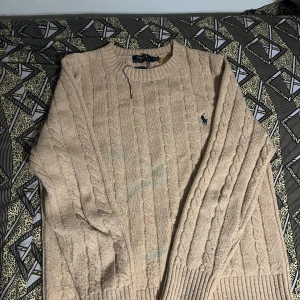 Beige stickad tröja från Ralph Lauren - Säljer en stilren beige stickad tröja från Ralph Lauren med kabelstickat mönster. Tröjan har långa ärmar och en rund halsringning. Den klassiska loggan är broderad på bröstet. Perfekt för kyligare dagar! Storlek M men passar även S