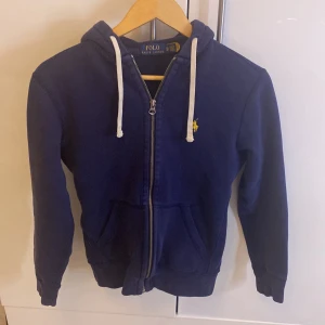 Mörkblå hoodie från Ralph Lauren stl XS - Säljer en snygg mörkblå hoodie från Ralph Lauren med dragkedja och vit snörning i huvan. Den har en broderad logga på bröstet och är perfekt för en stilren look. Passar bra till både jeans och joggers.