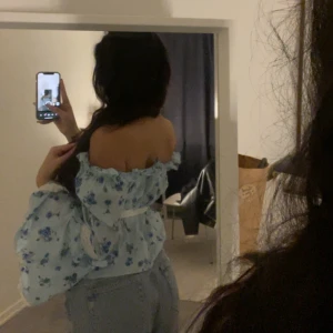 Oanvänd blommig babyblå sommar blus - Säljer en söt ljusblå offshoulder blus med blommigt mönster. Blusen har långa ärmar med volangdetaljer och spetskant. Den är knytbar framtill och har en luftig passform. Perfekt för en somrig look! 🌸