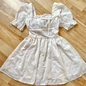 Vit klänning med puffärmar - Charmig vit klänning med korta puffärmar och broderade detaljer. Klänningen har en söt, romantisk stil med en smockad rygg för extra komfort. Perfekt för somriga dagar och speciella tillfällen.