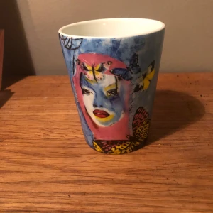 Mugg med konstnärligt motiv - Unik mugg med ett färgstarkt konstnärligt motiv av ett ansikte omgivet av fjärilar. Muggen har en blå bakgrund med inslag av rosa, gult och svart. Perfekt för att ge en konstnärlig touch till din morgonkaffe.