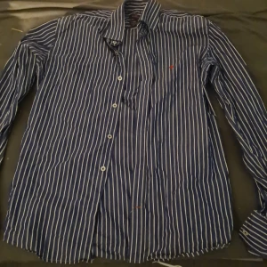 Morris skjorta - Snygg blå och vit randig skjorta från Morris med klassisk krage och knappar framtill. Den har en liten röd broderad logga på bröstet. Perfekt för en stilren look!