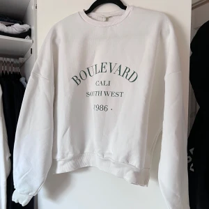 Vit sweatshirt med tryck - Säljer en vit sweatshirt med texten 'Boulevard Cali South West 1986' tryckt på framsidan. Tröjan är oversized i storleken, har långa ärmar och ribbade muddar vid ärmslut och nederkant. Perfekt för en avslappnad stil. Önskar du veta exakta mått? Tveka inte att höra av dig!