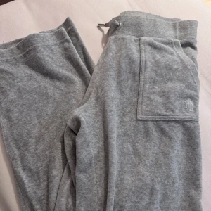 Grå mjukisbyxor från Juicy Couture - Säljer ett par grå mjukisbyxor från Juicy Couture. Byxorna har en bekväm passform med elastisk midja och snörning. De har praktiska fickor med knappar och en broderad logga på sidan. Perfekta för en avslappnad stil.